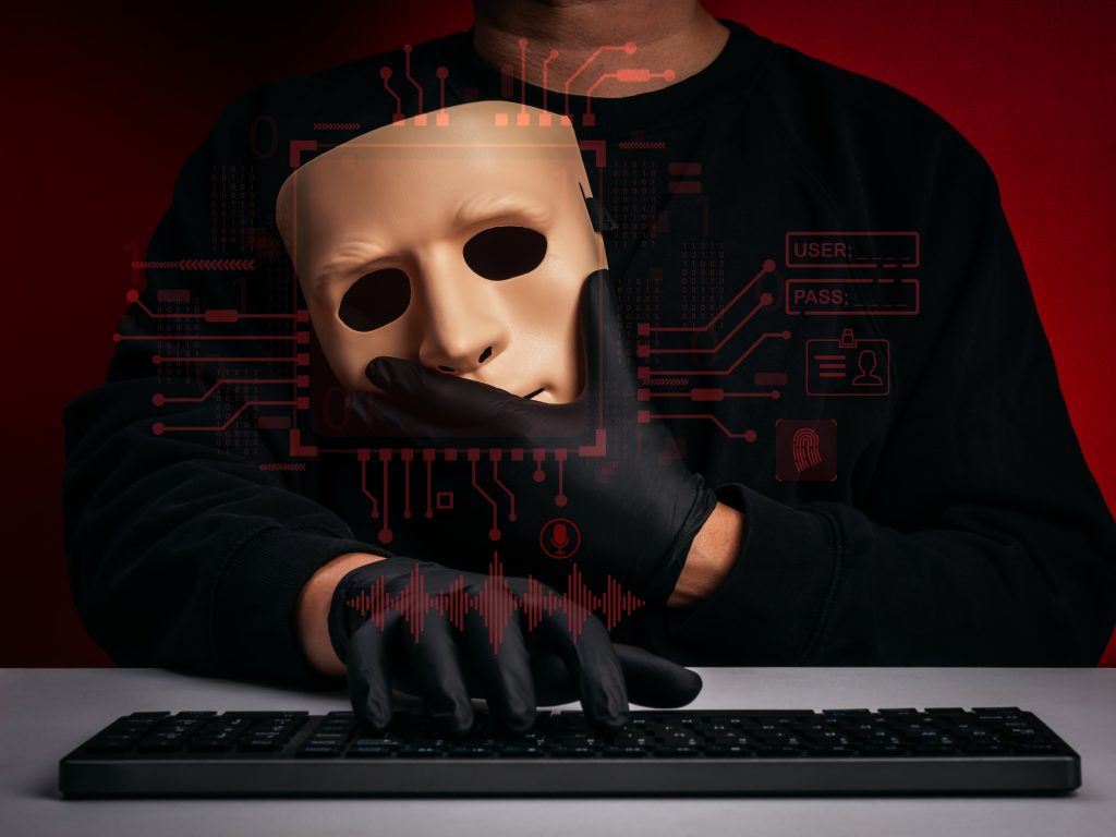 Header image: Black-clad hacker, shown from the neck down, on a dark red background, holding a humanlike mask in one gloved hand, while the other gloved hand rests on a keyboard (Credit: tete_escape / Shutterstock.com)